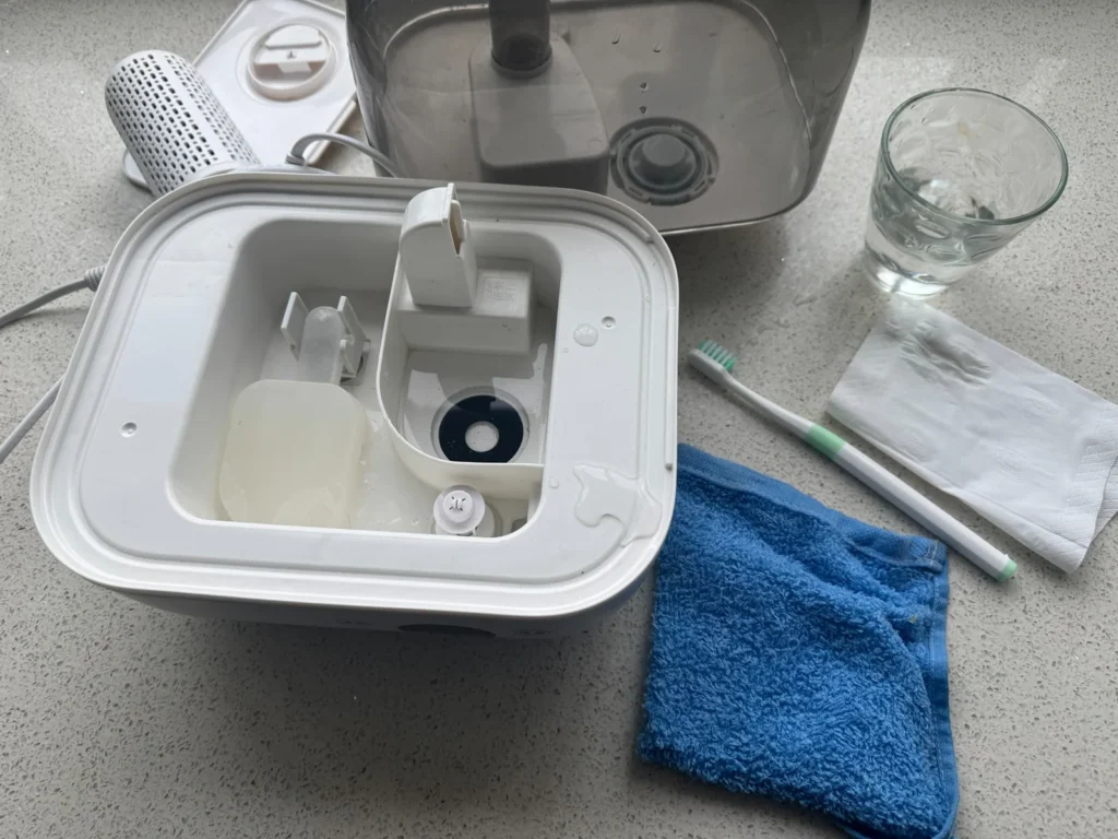 Open humidifier base with water tank, filter, toothbrush, cloth, and paper towels before cleaning