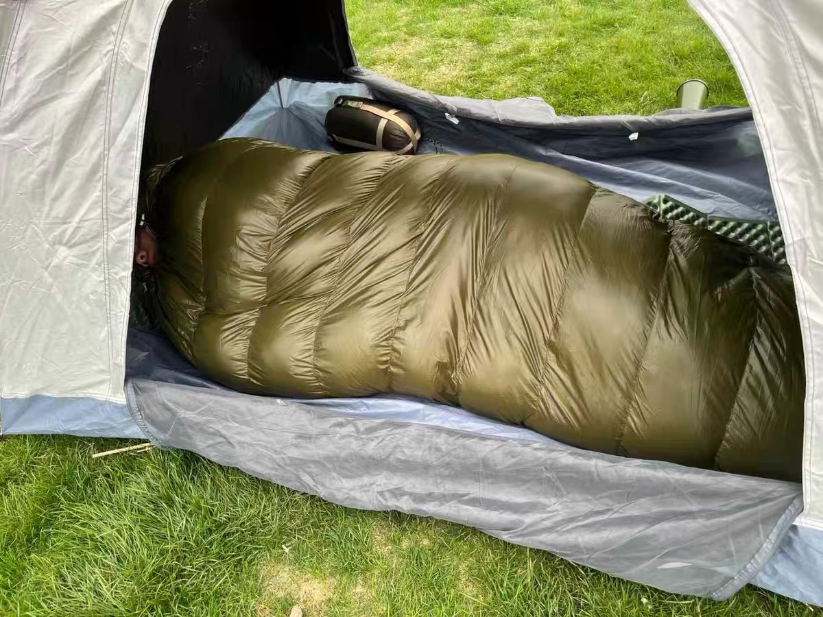 Olive-green sleeping bag inside an open gray tent at a grassy campsite.
