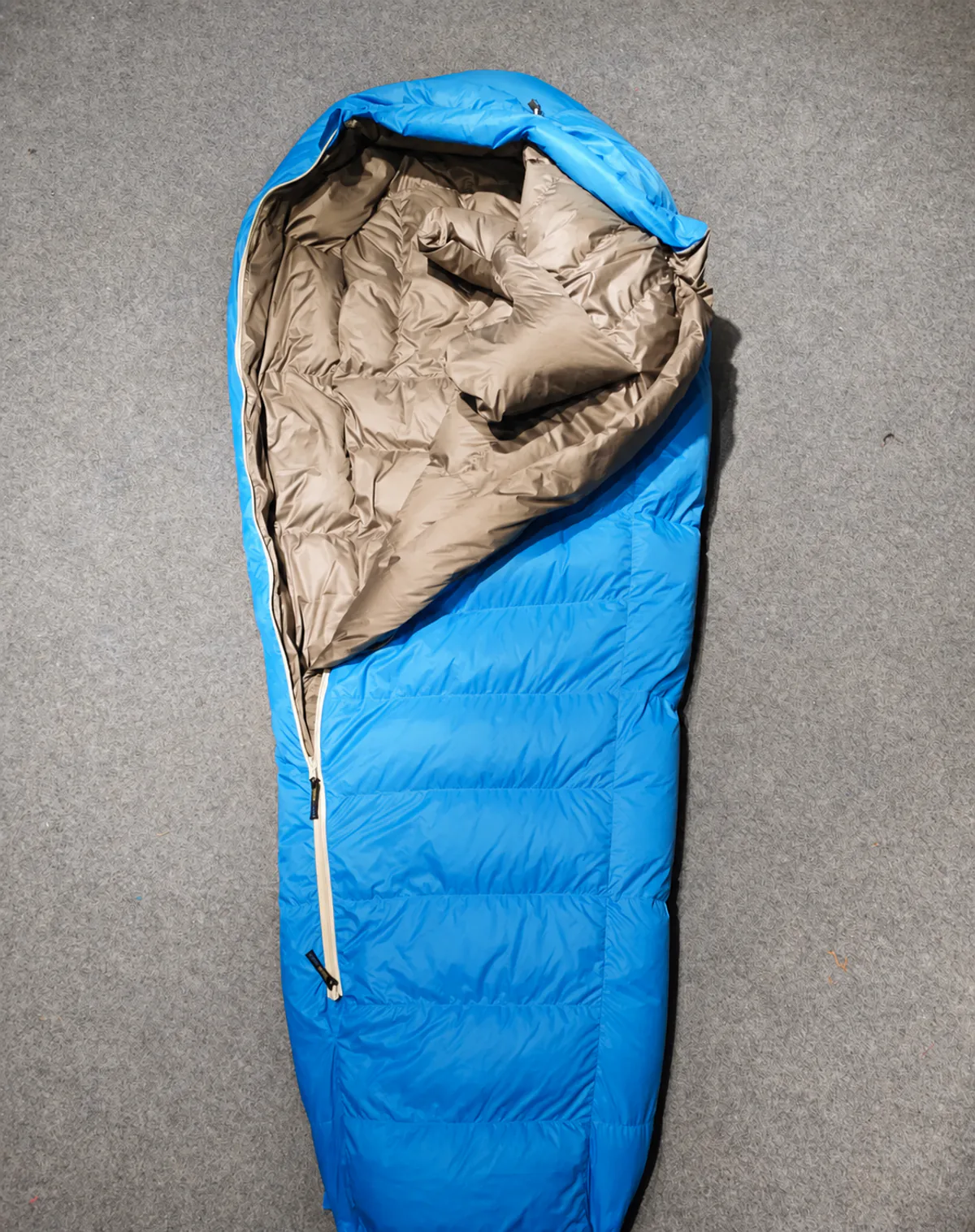 Blue mummy sleeping bag partially unzipped on a gray floor, showing the hood, side zipper, and beige lining.