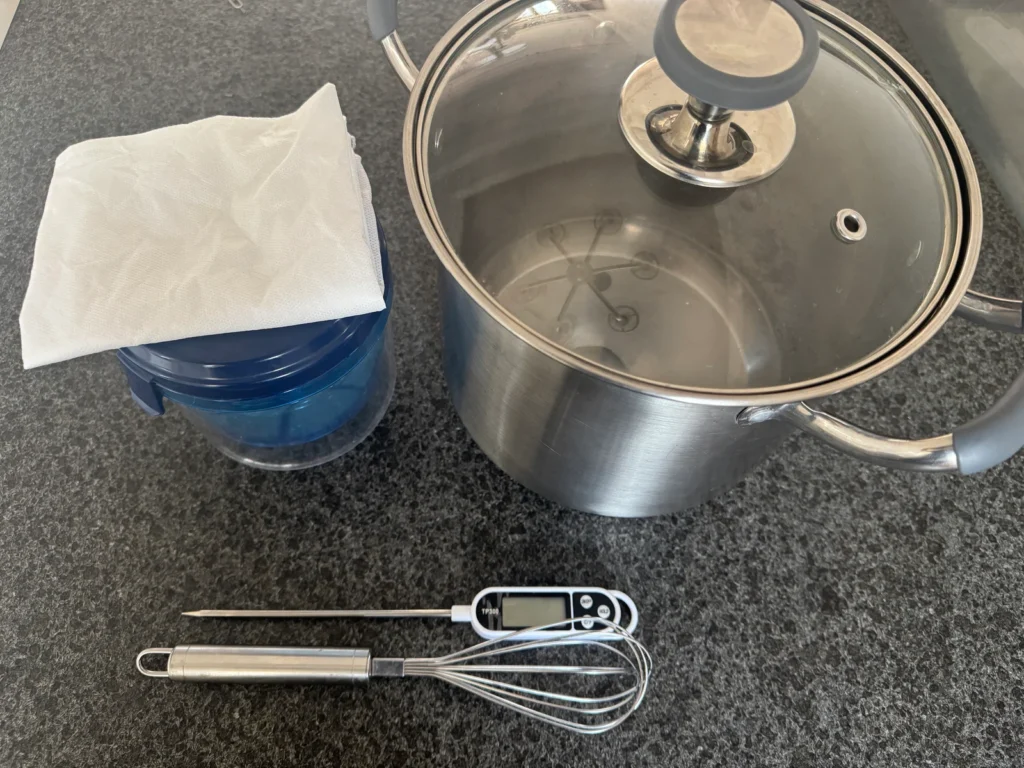 A stainless steel pot with a lid, a whisk, a digital thermometer, a blue yogurt strainer, and a white cloth.