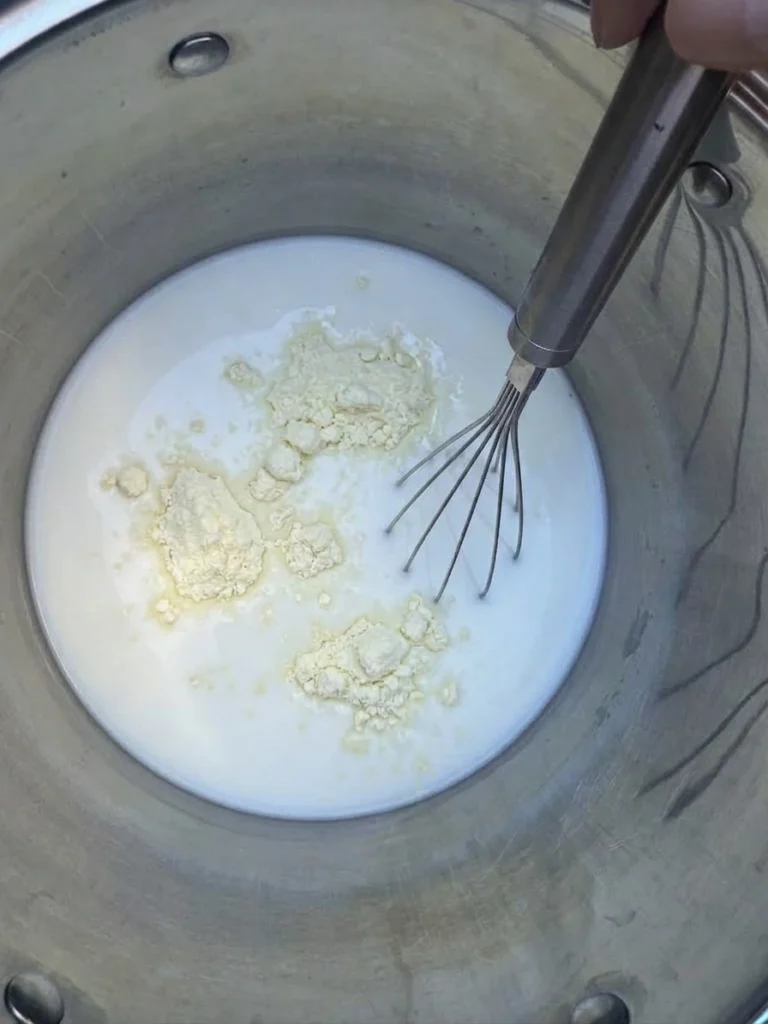 Whole milk powder sitting on the surface of milk beside a whisk in a stainless steel pot