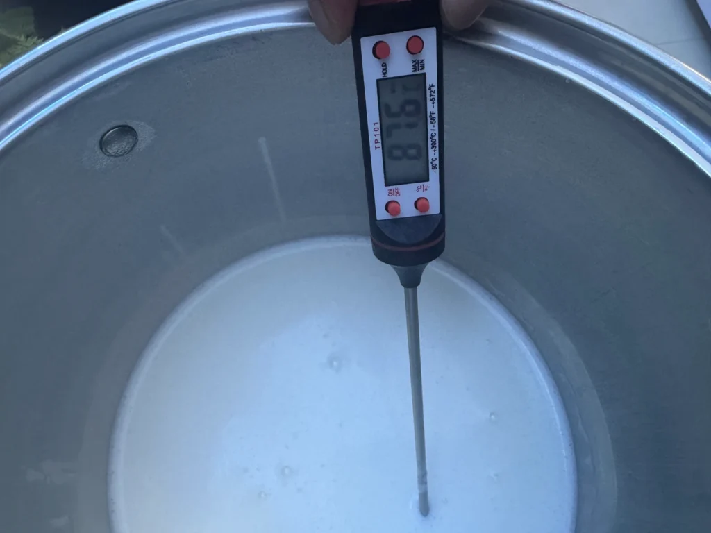 A digital thermometer reading about 87.6°C in milk inside a stainless steel pot.