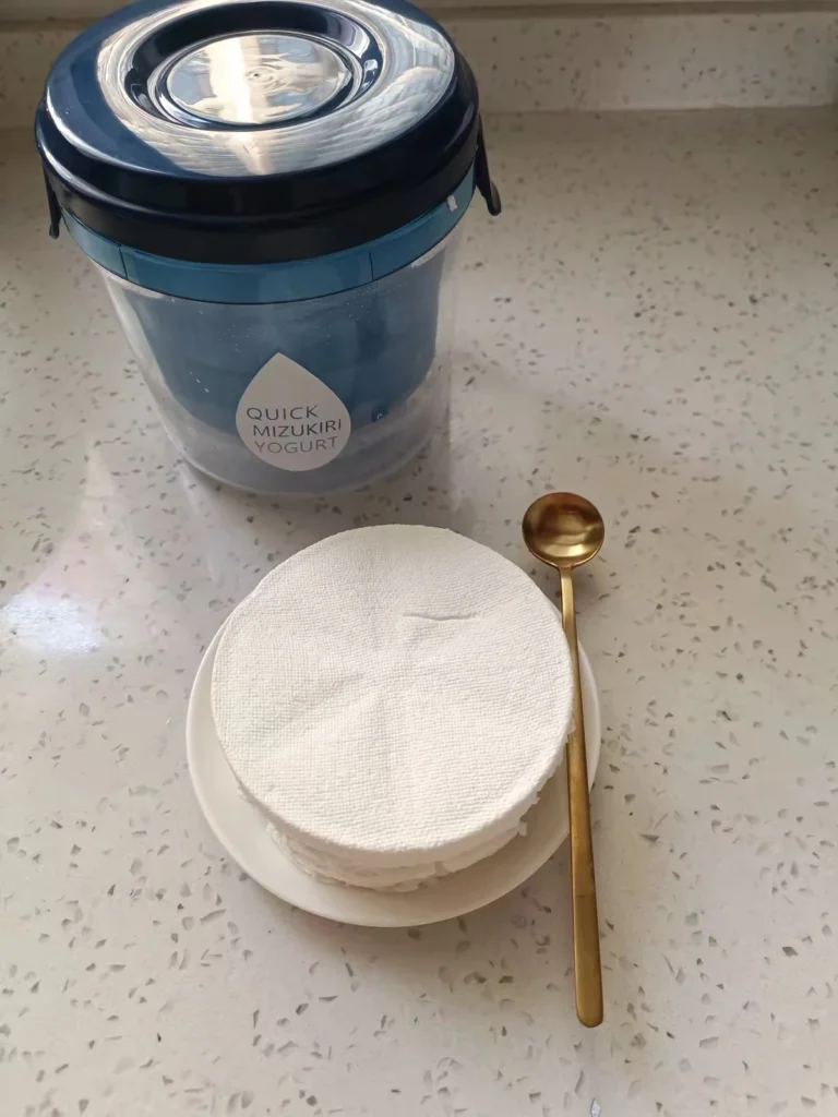 A blue yogurt strainer beside thick strained yogurt on a small white dish and a gold spoon.