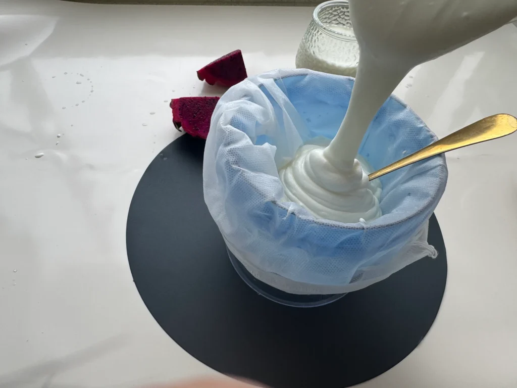 Yogurt being poured into a cloth-lined blue strainer with a gold spoon inside.