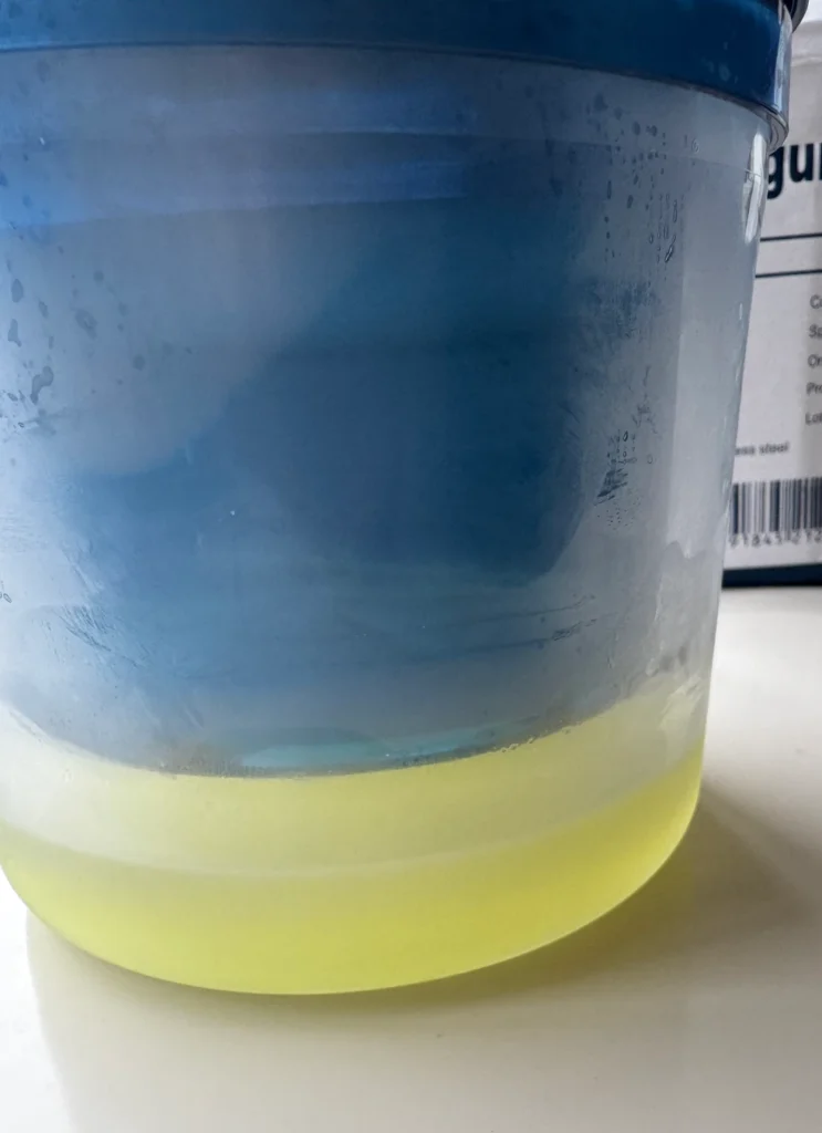 Pale-yellow whey collected at the bottom of a clear container beneath a blue yogurt strainer.