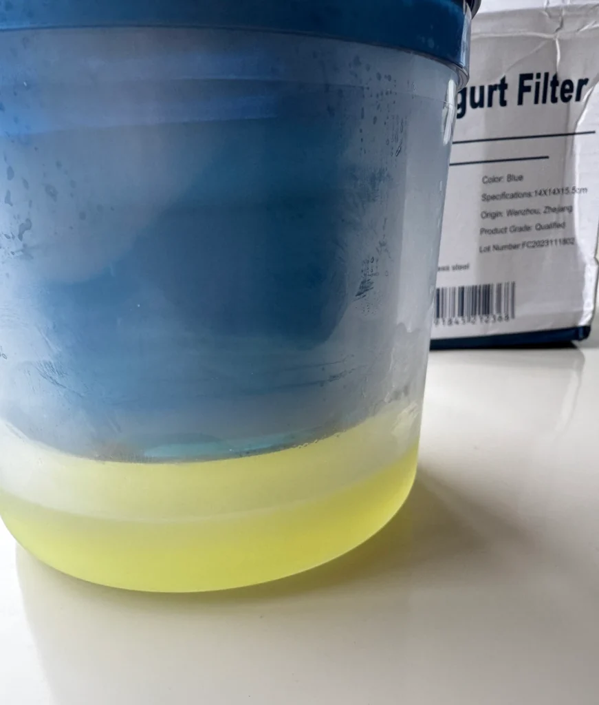 Pale-yellow whey collected at the bottom of a clear container beneath a blue yogurt strainer.