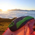 A green and dark sleeping bag draped over an orange tent at sunrise above a sea of clouds.
