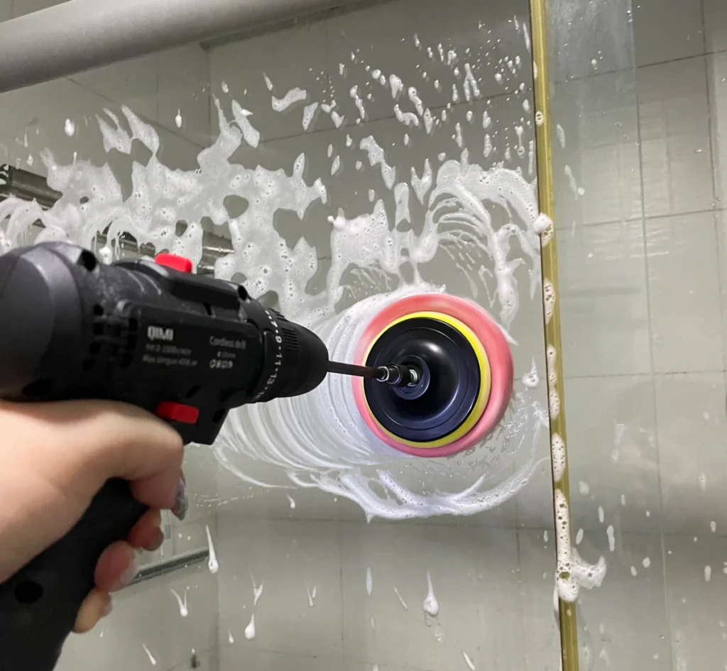 A hand using a drill-mounted circular cleaning pad on a soap-covered shower glass panel.