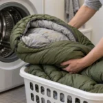A person handling a used olive-green sleeping bag in a laundry basket beside a front-loading washing machine.
