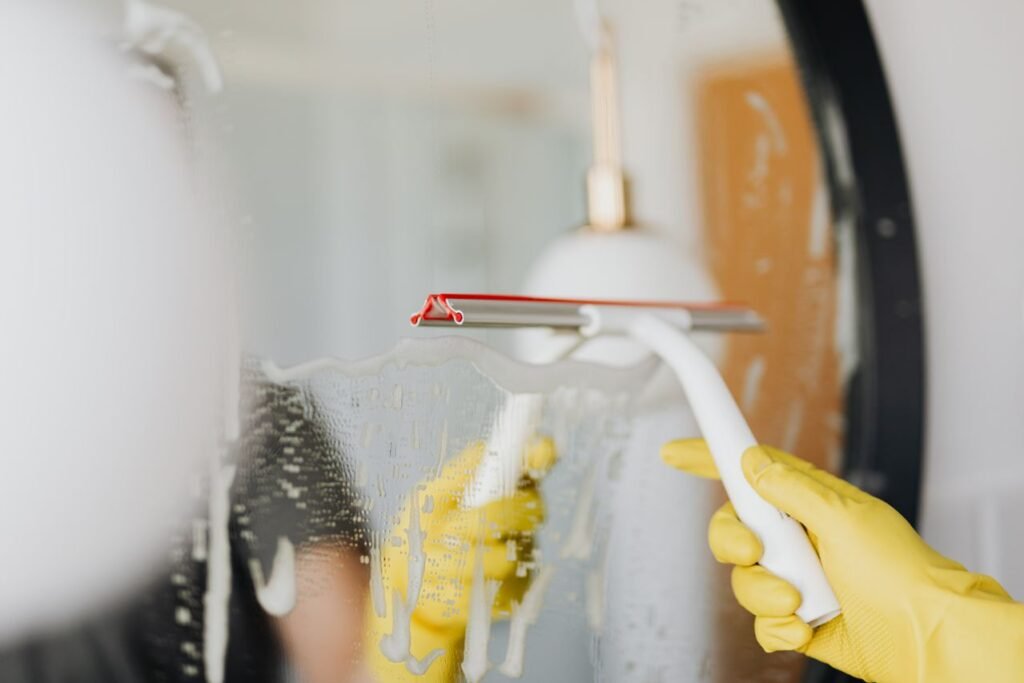 A yellow-gloved hand using a squeegee to remove soapy water from a bathroom mirror.